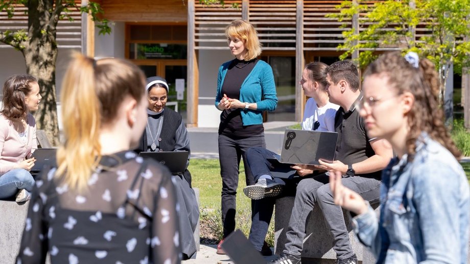 Sechs Studierende sitzen auf Steinblöcken im Kreis und reden miteínander. In der Mitte steht eine Dozentin. Drumherum ist Natur, im Hintergrund ein Audimax-Gebäude.
