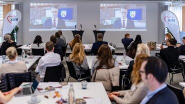 Eine Gruppe von Personen nimmt in einem Konferenzraum an einer Videokonferenz teil. Auf zwei großen Bildschirmen ist der NRW-Ministerpräseident Wüst in Nahaufnahme zu sehen, der virtuell zugeschaltet ist. Die Teilnehmer sitzen an langen Tischen und verfolgen aufmerksam die Präsentation. Im Raum sind zwei Podeste mit Mikrofonen sowie Banner mit dem Logo 'AWO' zu erkennen.