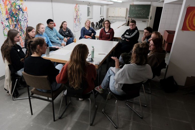 Ca. 15 Studierende sitzen um einen Tisch in einem Kellerraum. Sie hören der Person im Vordergrunmd zu. An den Wänden sind selbstgemalte Bilder aufgehangen.