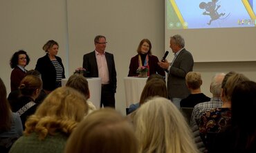 Drei Frauen und zwei Männer stehen an Tischen, eine der Männer spricht in ein Mikrofon. Im Vordergrund ist das Publikum.