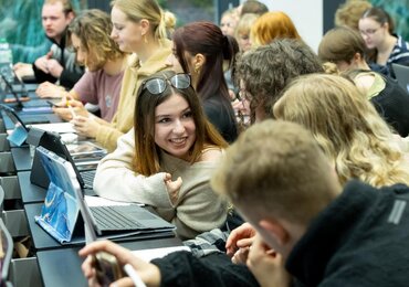 Studierende sitzen in einem Hörsaal und sind lächelnd in ein Gespräch vertieft.