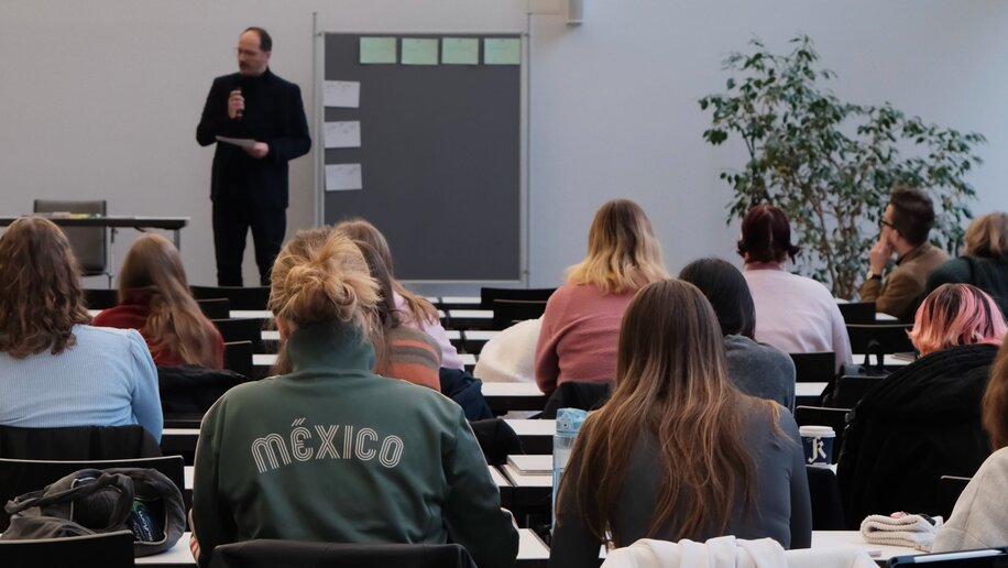 Ein Dozent spricht in ein Mikrofon, neben ihm eine Pinnwand. Im Vordergrund sitzen Studierende in Tischreihen.