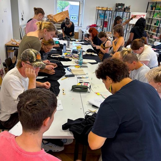 Studierende sitzen zusammen an einem Tisch und haben vor sich schwarze T-Shirts und Stickrahmen. Sie besticken die T-Shirts mit weißem Faden.