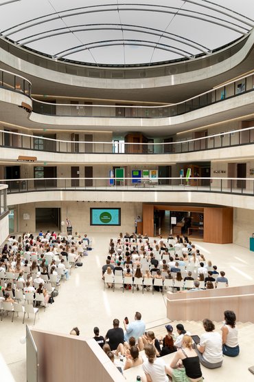 Innenbereich eines modernen Gebäudes mit Stuhlreihen und Treppenstufen. Auf den Stühlen und Treppenstufen sitzen Menschen. 