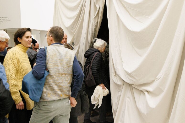 Eine Gruppe Menschen geht durch einen weißen Vorhang, in das innere einer Höhlen artigen Konstruktion.