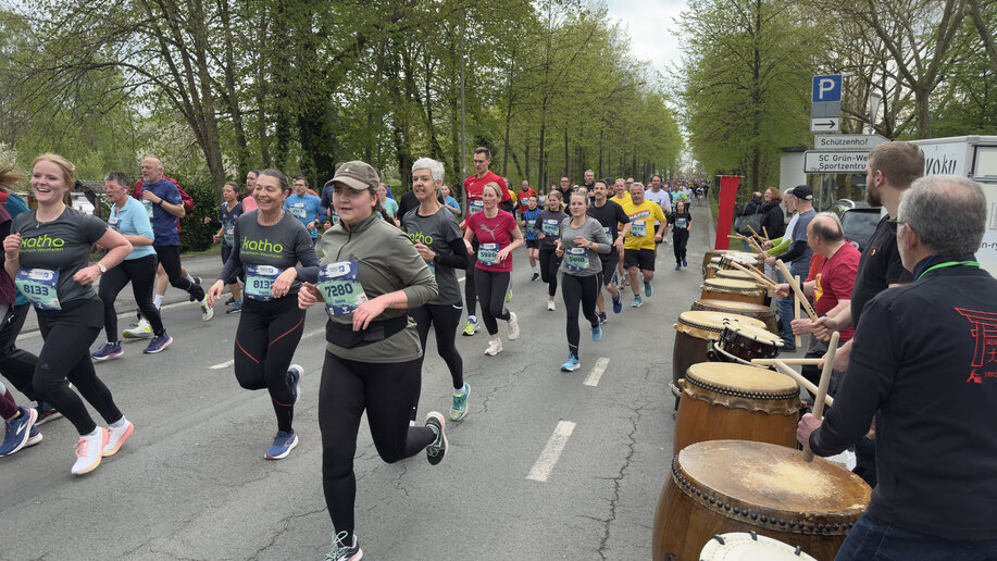 Einige Läufer_innen des katho-Teams auf der Rennstrecke. Am Straßenrand eine anfeuernde Musikgruppe mit Trommeln.