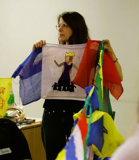 Professorin Ziese hält eine Schnur mit bunten Flaggen und blickt zur Seite. Auf der Flagge vor ihr ist eine Frau mit einer Krone gemalt, zu deren Füßen stehen kleine schwarze Figuren.