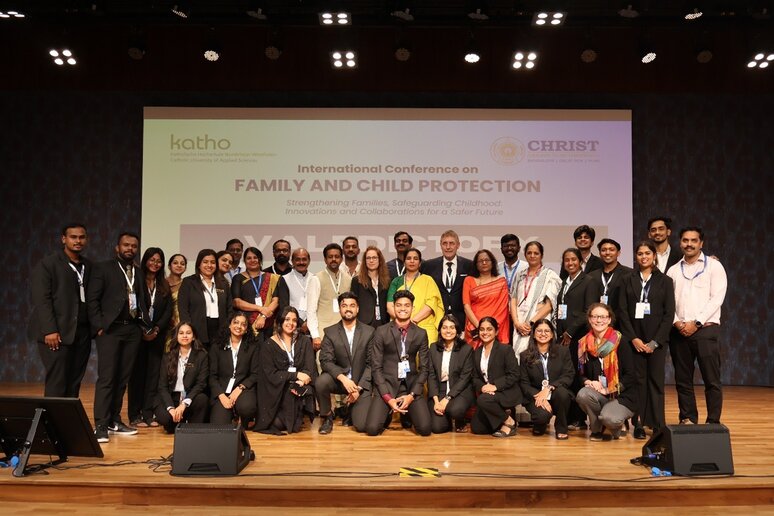 Eine große Gruppe von Menschen posiert auf einer Bühne vor einer Leinwand, die eine internationale Konferenz über Familien- und Kinderschutz ankündigt. Die Personen sind größtenteils formell gekleidet und tragen Namensschilder. Der Anlass scheint Teil einer feierlichen Veranstaltung zu sein. Die Atmosphäre wirkt professionell und engagiert.