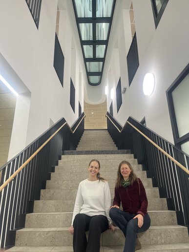 Frau Labenz und Frau Schäfer-Wiesen sitzen auf einer Treppe in der Hochschule Campus Wien und blicken in die Kamera.