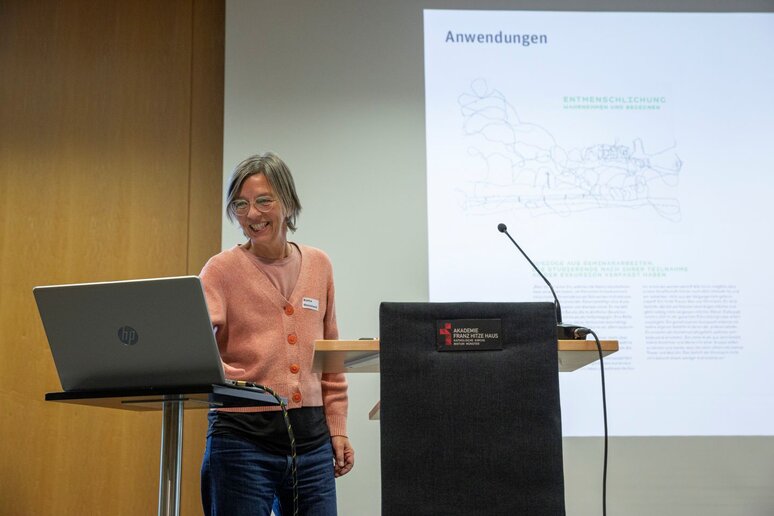 Eine Frau mit grauen Haaren und oranger Strickjacke steht auf der Bühne vor einem Rednerpult. Sie lacht und gibt dabei etwas am Laptop ein.