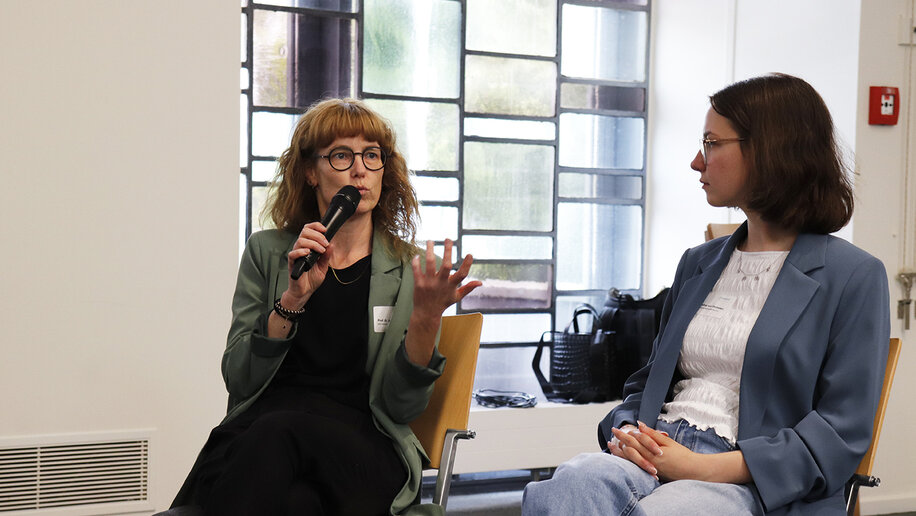 Zwei Frauen bei einer Podiumsdiskussion. Die linke Frau hält ein Mikrofon und spricht, die rechts hört zu.