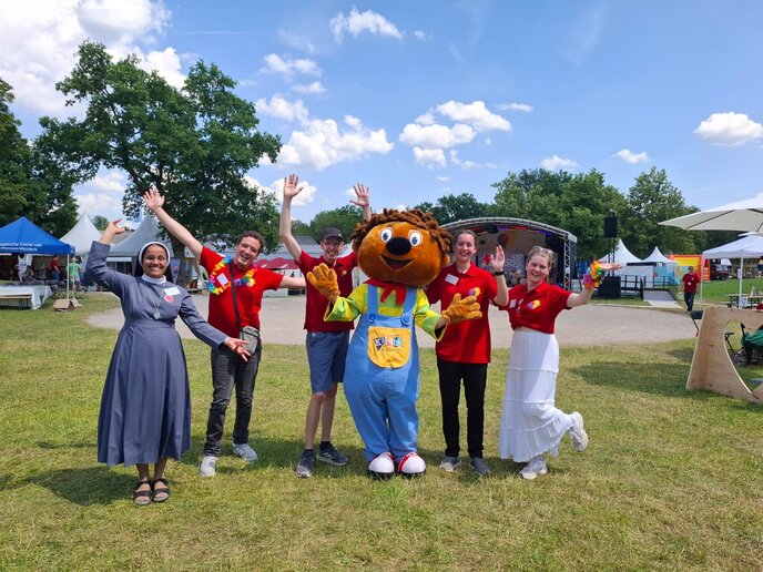 Ein lebensgroßes Maskottchen mit rotem Kopf und blauer Hose steht zusammen mit fünf Studierenden auf einer Wiese. Alle schauen fröhlich.