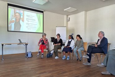 Mehrere PErsonen, darunter dioe Ordensschwetsr und der Professor sitzen im Halbkreis im Seminarraum. An der Wand sind Foto und Infos zu Schwester Nirina projiziert.-