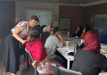 Professorin spricht mit einer Teilnehmerin zu ihr heruntergebeugt. Die Teilnehmerin sitzt mit dem Rücken zur Kamera und schaut Pofessorin an. Um sie herum sitezn weitere Teilnehmerinnen an Tischen.