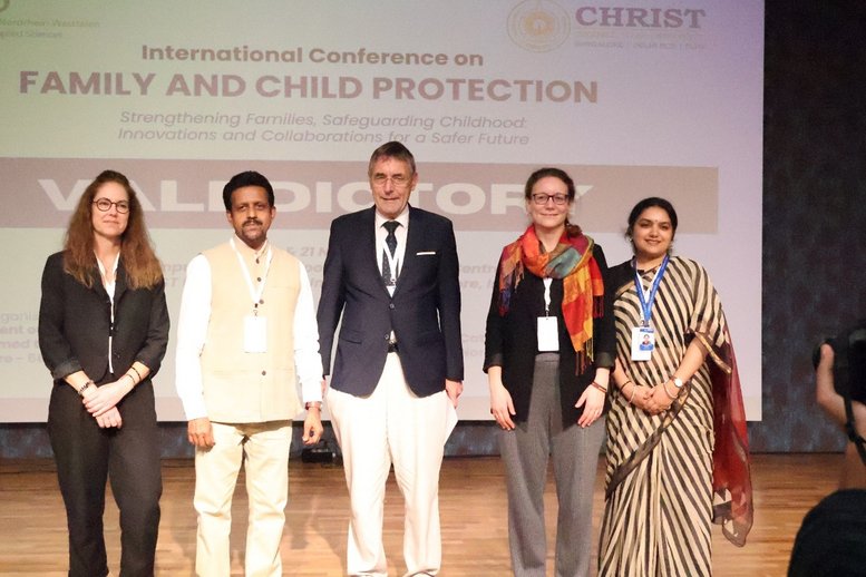 Fünf Personen stehen auf einer Bühne vor einer Leinwand, die eine internationale Konferenz über Familien- und Kinderschutz ankündigt. Sie sind formell gekleidet und tragen Namensschilder, was auf ihre Teilnahme an einer bedeutenden Veranstaltung hinweist. Die Atmosphäre wirkt professionell und engagiert.