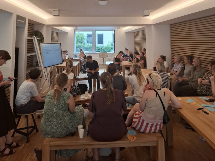 Menschn sitzen in Gruppen verteilt auf Bänkden der Cafeteria. In ihrer Mitte befinden sich ein Lehrender und ein digitales Whiteboard.
