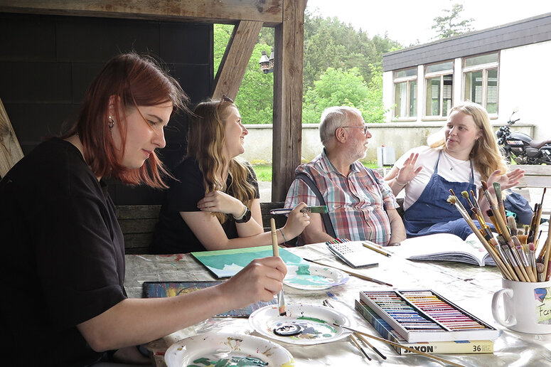 Vier Menschen sitzen zusammen am Tisch. Drei unterhalten sich und eine malt.
