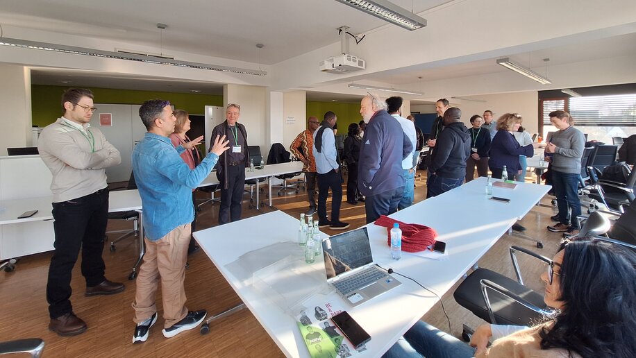 Menschen stehen in kleinen Gruppen in einem Konferenzraum und unterhalten sich angeregt. Einige gestikulieren, andere hören aufmerksam zu. Laptops und Getränkeflaschen befinden sich auf den Tischen. Der Raum ist modern und lichtdurchflutet, mit entspanntem und lebendigem Ambiente.