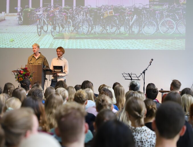 Zwei Vertreter aus AStA und StuPa begrüßen die neuen Studierenden am Pult auf der Bühne.