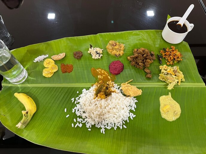 Kerala Sadya: Weiße Reisportion und zahlreiche kleine vegetarische Beilagen auf einem großen Bananenblatt; daneben eine Banane, ein Glas Wasser und eine Tasse.
