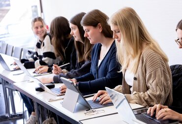 Mehrere Personen sitzen hintereinander an einem Tisch an ihren Laptops und arbeiten.
