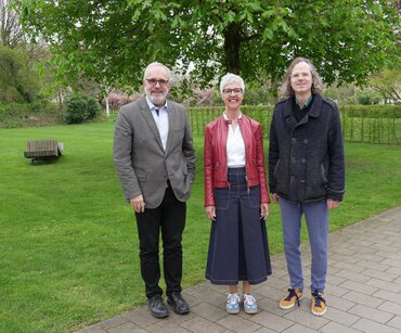 Gruppe von zwei Männern und einer Frau (Mitte), die auf einem Weg vor einer grünen Wiese stehen und in die Kamera lächeln.