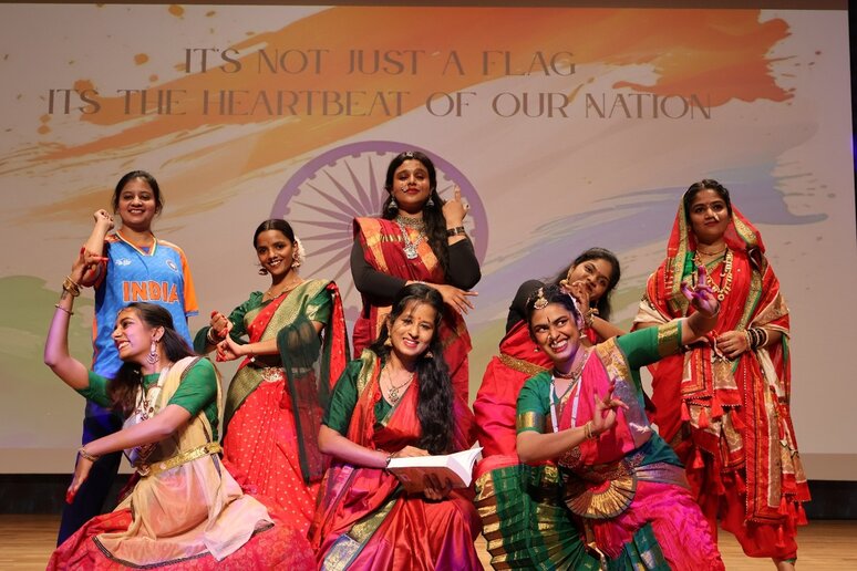 Eine Gruppe von Frauen posiert in traditionellen indischen Kleidern auf einer Bühne. Eine Frau trägt ein Trikot mit der Aufschrift "India". Im Hintergrund ist eine stilisierte indische Flagge und der Schriftzug "It's not just a flag, it's the heartbeat of our nation" zu sehen. Die Szene wirkt festlich und feierlich.