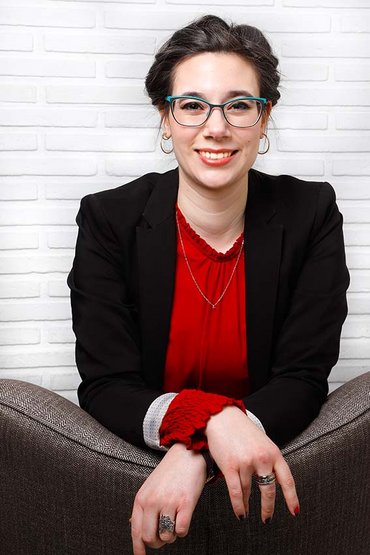 weiblich gelesene Person kniet auf einem Sessel, die Arme liegen überkreuzt auf der Rückenlehne, vor einer weißen Wand und lächelt in die Kamera. Ihre Haare sind zusammengebunden, sie trägt eine dunkelgrüne, rechteckige Brille, ein rotes Oberteil und einen schwarzen Blazer darüber. 