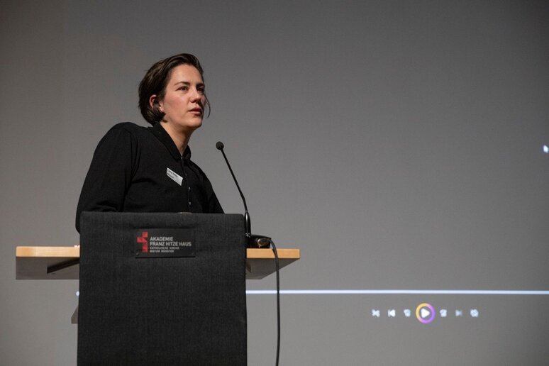 Eine Frau steht am Rednerpult. Sie hat kurze Kinnlange Haare und trägt schwarze Kleidung.