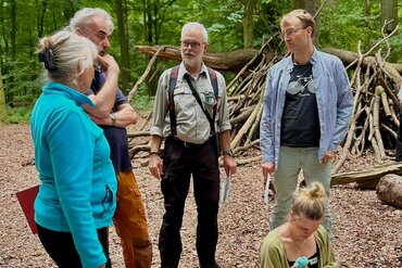 Vier Personen stehen im Halbkreis im Wald vor aufgestapeltem Holz. Eine junge Frau sitzt davor.