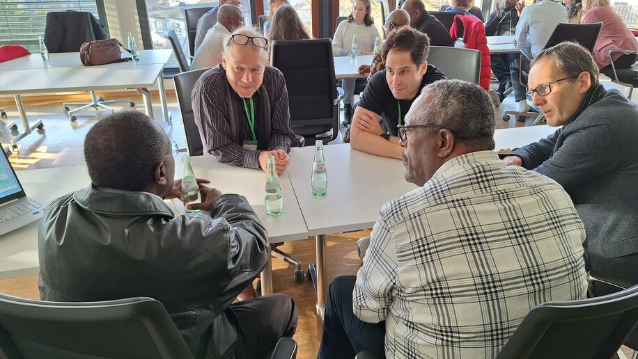 Mehrere Personen sitzen in einer kleinen Gruppe an einem Tisch und diskutieren miteinander. Jeder hat eine Wasserflasche vor sich, und sie wirken konzentriert und engagiert. Im Hintergrund sind weitere Gruppen zu sehen, die ebenfalls in Gespräche vertieft sind. Der Raum ist hell, mit großen Fenstern, die viel Licht hereinlassen.