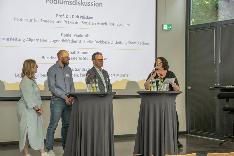 Vier Redner_innen stehen an Stehtischen auf der Bühne und diskutieren