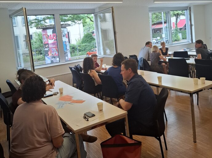 Ca. 10 Menschen sitzen in Gruppen an drei Seminartischen.