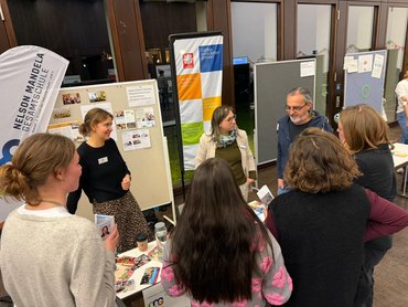 6 Personen stehen rund um einen Tisch und unterhalten sich.
