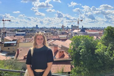 Eine junge Frau schaut in die Kamera. Im Hintergrund sieht man das Stadtpanorama von Zagreb vor blauem Himmel.