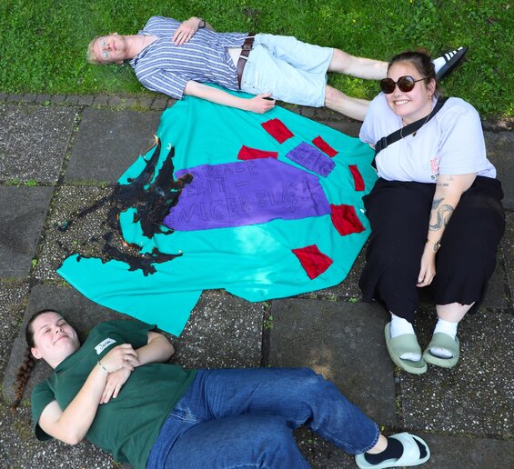 Zwei Studentinnen und ein Student liegen bzw sitzen auf einem Steinweg neben einer Wiese und lächeln. In ihre Mitte ist ein großer grüner Stoff mit roten Aufnähern und einem Schriftzig auf lila stoff: "Mehr Soziale Arbeit - weniger Bla". An einem Teil des Stoffes sind kleine Flammen sichtbar, der Schriftzug ist angebrannt.