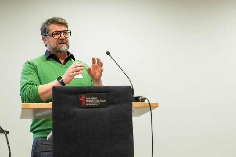 Ein Mann steht vor einem Rednerpult. Er trägt einen grünen Pullover, hat dunkel-graue Haare und eine große schwarze Brille.