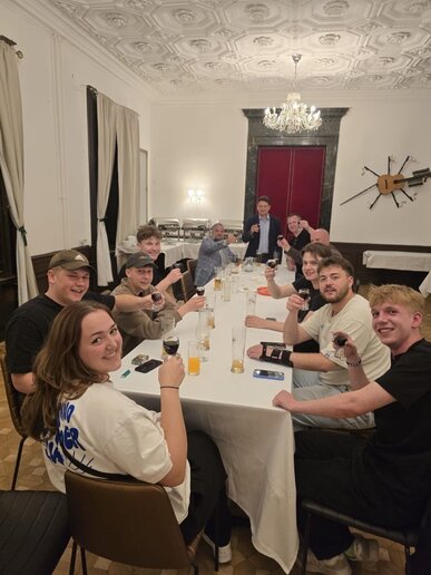Eine Studierendengruppe sitzt an einem Tisch in einem Restaurant und halten ihre Getränke in die Kamera.