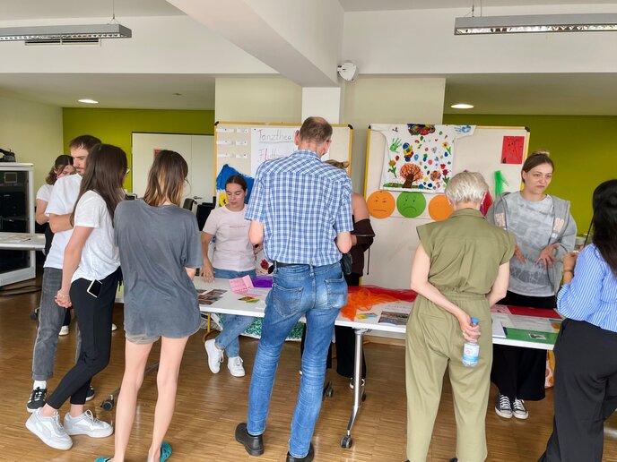 Interessierte Teilnehmer_innen stehen vor dem Stand einer Projektgruppe und betrachten die ausgelegten Gegenstände wie Fotos.