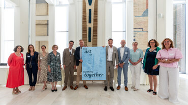 Elf Männer und Frauen in festlicher Kleidung stehen in einer Reihe. Zwei Männer in der Mitte halten ein großes Plakat mit der Aufschrift "The art of working together".