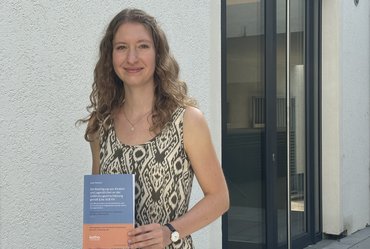 Person steht vor einer weißen Wand mit brauner Tür, blickt freundlich in die Kamera und hält ein Buch mit beiden Händen vor dem Bauch. 
