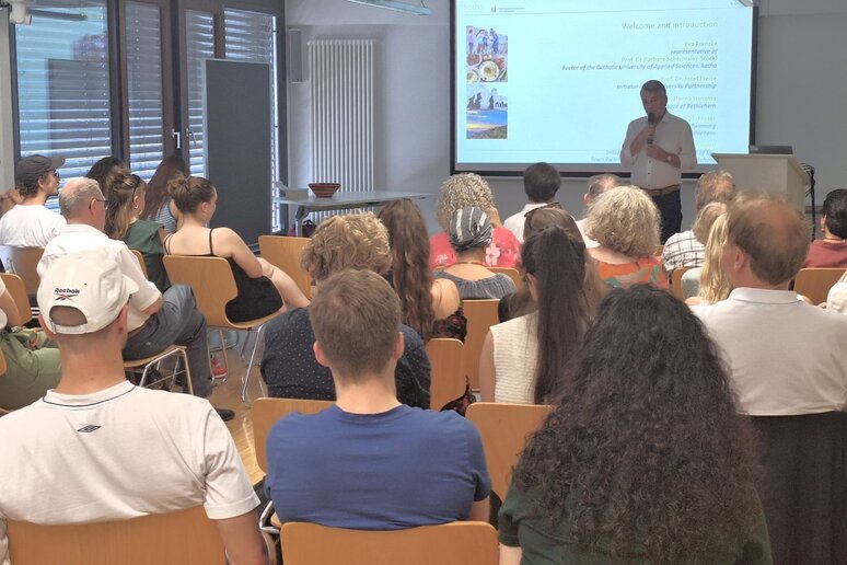 Vor mehreren mit Personen besetzten Sitzreihen steht ein Mann mit Mikrofon und spricht