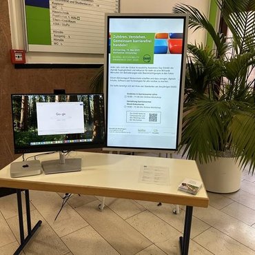 Im Foyer steht ein Tisch, auf dem sich ein Bildschirm, Infozettel und Postkarten befinden. Dahinter steht ein digitaler Bildchirm im Hochkantformat, auf diesem ist werbung für den GAAD-Tag an der katho zu sehen.