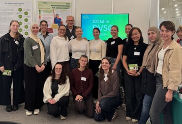 Ein Gruppenfoto mit 12 Studierenden und 3 Lehrenden, die vor einem Bildschirm mit der Aufschrift 100 Jahre DVSG stehen. Im Hintergrund sind Poster über Wissenschaftsprojekte zu sehen