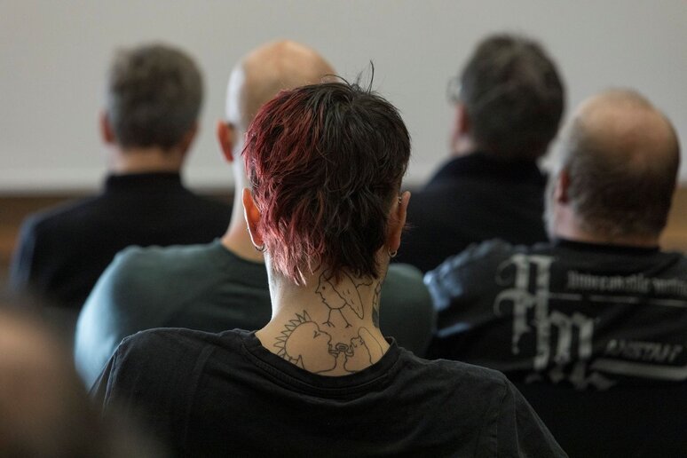 Eine Frau mit kurzen roten Haaren sitzt in einer Stuhlreihe und wird von hinten fotografiert. Es ist nur ihr Kopf und tätowierter Nacken zu sehen.