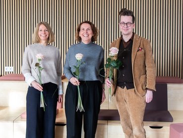 Drei Personen (zwei Frauen und ein Mann) stehen mit einer Rose lächelnd vor einer Wand. 