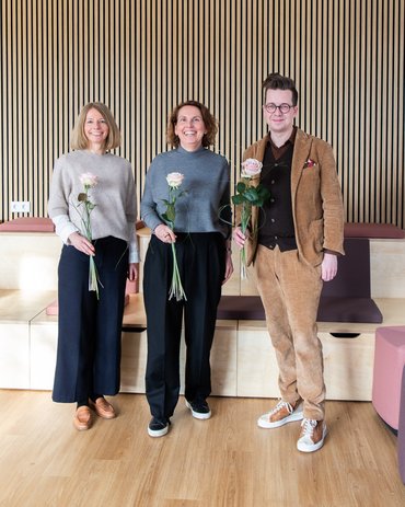 Drei Personen (zwei Frauen und ein Mann) stehen mit einer Rose lächelnd vor einer Wand. 