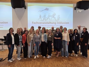 Eine lächelnde Gruppe von Menschen steht in einem Vorlesungssaal vor einer Leinwand. Alle Personen freuen sich sehr auf den Fachtag. 