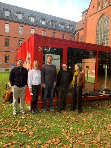 Fünf Personen stehen vor einem roten Container, der als Ausstellungsraum dient. 