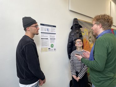 Drei Personen stehen vor einem Poster und reden miteinander.